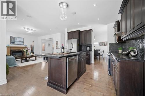 Gourmet kitchen featuring dark wood cabinetry, granite countertops, stainless steel appliances, a mosaic tile backsplash, and a central island with seating, opening to a living area with a fireplace - 145 Redtail Street, Kitchener, ON - Indoor Photo Showing Kitchen With Stainless Steel Kitchen With Upgraded Kitchen