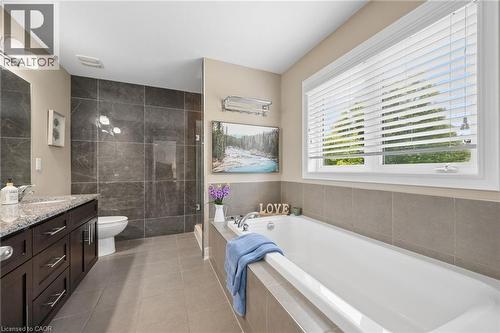 Spacious bathroom featuring a large soaking tub, separate glass-enclosed shower with dark stone-finish tiling, a vanity with ample storage, a stone-finish countertop, and a window with blinds - 145 Redtail Street, Kitchener, ON - Indoor Photo Showing Bathroom
