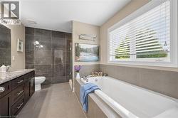 Spacious bathroom featuring a large soaking tub, separate glass-enclosed shower with dark stone-finish tiling, a vanity with ample storage, a stone-finish countertop, and a window with blinds - 