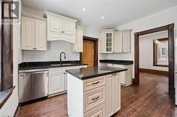 Kitchen featuring a central island with a dark countertop, white cabinetry with dark hardware, a stainless steel dishwasher, a white subway tile backsplash, and dark wood-finish flooring - 