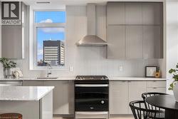 Modern kitchen featuring grey flat-panel cabinetry, a stainless steel range hood, white subway tile backsplash, and light-colored countertops - 