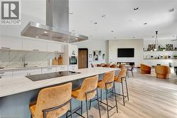 Spacious open-concept living area featuring a kitchen island with built-in cooktop, stainless steel range hood, white cabinetry with stone-finish backsplash, and light wood-finish flooring - 
