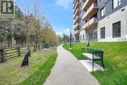 Modern multi-story building featuring a gravel pathway, mature trees, a wooden split-rail fence, a green slatted bench, and a grass embankment - 