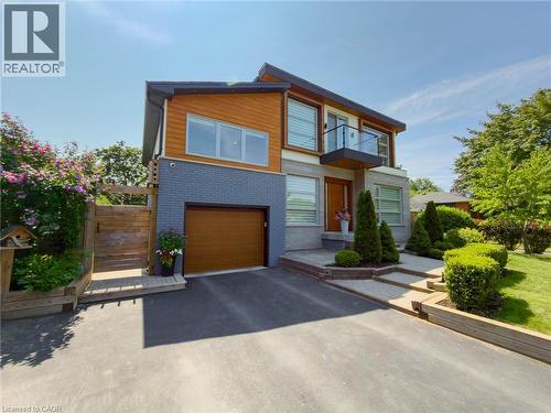 Modern exterior with a wood-finish garage door, grey brick and wood-finish siding, a second-story balcony with glass railing, a paved driveway, and established front landscaping - 5178 Hunter Drive, Burlington, ON - Outdoor