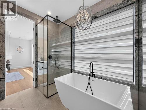 Freestanding soaking tub with a matte black faucet, glass-enclosed shower, large format tile flooring, stone-clad walls, and a decorative crystal chandelier - 5178 Hunter Drive, Burlington, ON - Indoor