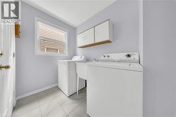 Dedicated laundry area featuring white tile flooring, upper cabinetry with wood-finish trim, a utility sink, and a window with blinds - 