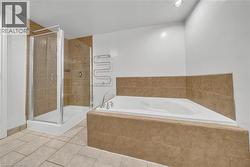 Spacious bathroom featuring a glass-enclosed shower with tile surround, an oversized built-in soaking tub with tile surround, a wall-mounted towel warmer, recessed lighting, and large format floor til - 