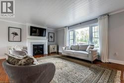 Living room featuring hardwood flooring, a fireplace with a stone surround and white mantel, a large multi-pane window, and a white plank ceiling - 