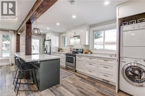 Modern kitchen featuring a central island with seating, stainless steel appliances, white cabinetry, wood-finish flooring, and a stacked washer/dryer unit - 323 Cranbrook Drive, Hamilton, ON - Indoor Photo Showing Laundry Room
