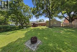 Expansive backyard featuring a large lawn area, mature trees providing natural shade, a built-in fire pit with a stone surround, a privacy fence, and a storage shed - 