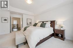 Carpeted bedroom featuring a tray ceiling, white walls, and an open doorway revealing a stairwell with dark wood flooring and a wooden banister - 