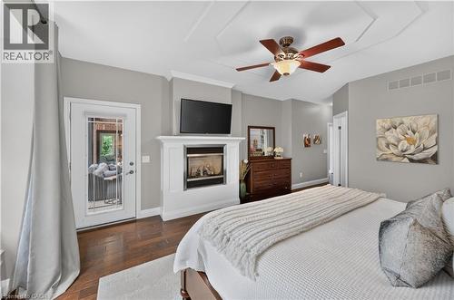 Spacious room featuring wood-finish flooring, a tray ceiling with a ceiling fan, a fireplace with a white mantel, and a glass-paned door - 2050 15 Side Road, Milton, ON - Indoor Photo Showing Bedroom With Fireplace
