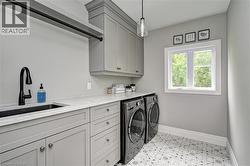 Laundry room featuring light gray cabinetry, a matte black faucet with an undermount sink, white countertops, pattern tile flooring, and a window providing natural light - 