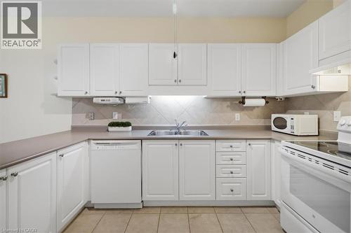 U-shaped kitchen featuring white cabinetry, light-toned countertops, a tile backsplash, and light beige floor tiles - 34 Southbrook Drive Unit# 13, Binbrook, ON - Indoor Photo Showing Kitchen With Double Sink