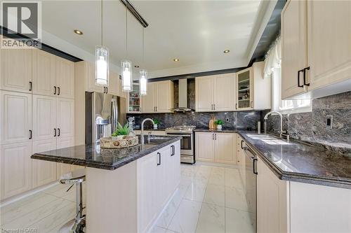 Contemporary kitchen featuring cream-toned cabinetry, dark stone countertops, stainless steel appliances, a center island with pendant lighting, and white tile flooring - 2380 Woodcrest Drive, Oakville, ON - Indoor Photo Showing Kitchen With Upgraded Kitchen