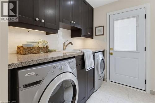 Laundry area featuring dark wood cabinetry, white subway tile backsplash, light-colored stone-finish countertops, an undermount sink with a chrome faucet, and tile flooring - 2188 Bader Crescent, Burlington, ON - Indoor Photo Showing Laundry Room