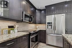 Contemporary kitchen featuring dark wood-finish cabinetry, light-toned stone countertops, a tiled backsplash, stainless steel appliances, and integrated under-cabinet lighting - 