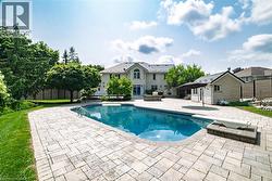 Expansive backyard featuring a large swimming pool with a diving board, surrounding paver patio, a stone-clad pool house with a dark-tiled roof, and mature perimeter trees - 