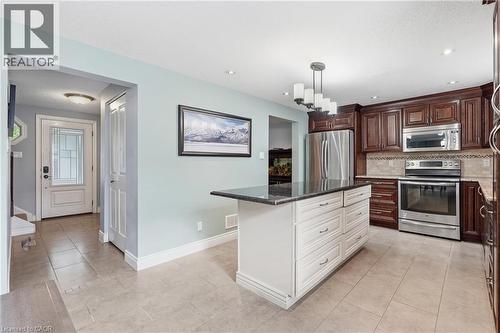 Kitchen featuring a central island with white cabinetry and dark countertop, complemented by rich wood-finish upper and lower cabinetry, stainless steel appliances, and a tiled backsplash - 19 Alona Avenue, Cambridge, ON - Indoor Photo Showing Kitchen