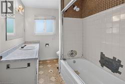 Bathroom featuring a white vanity with an integrated sink and mirror, a window with blinds, a toilet, and a bathtub with white subway tile surround and a brown mosaic tile accent - 