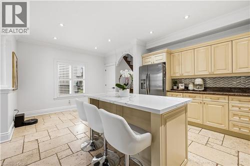 Kitchen featuring a central island with a white countertop, beige cabinetry, stainless steel refrigerator, patterned tile backsplash, and light-toned tile flooring - 199 Hunter Street W, Hamilton, ON - Indoor Photo Showing Kitchen