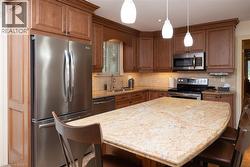 Kitchen featuring wood cabinetry, stainless steel appliances, a corner sink with a window, and a light-toned granite island with pendant lighting - 
