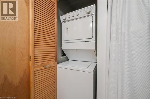 White stackable washer and dryer unit, featuring a top-loading washer and front-loading dryer, encased by louvered bi-fold doors with a wood-finish - 26 Ontario Street Unit# 127, Guelph, ON - Indoor Photo Showing Laundry Room