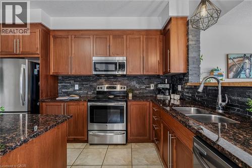 Kitchen featuring wood cabinetry, stainless steel appliances, dark stone countertops, and a stacked stone tile backsplash - 174 Woodway Trail, Simcoe, ON - Indoor Photo Showing Kitchen With Double Sink