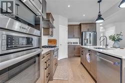 Modern kitchen featuring stainless steel appliances, a center island with sink, pendant lighting, wood-finish flooring, and shaker-style cabinetry - 