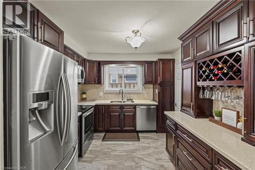 Kitchen featuring dark wood cabinetry, stainless steel appliances, light-toned countertops, a tile backsplash, and a built-in wine rack with stemware storage - 167 Berkindale Drive, Hamilton, ON - Indoor Photo Showing Kitchen With Double Sink