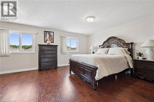 Spacious primary bedroom featuring rich wood-finish flooring, ornate dark wood bed frame with decorative headboard, dual nightstands with attached lighting, and ample natural light from two large wind - 86 Bridlewreath Street, Kitchener, ON - Indoor Photo Showing Bedroom