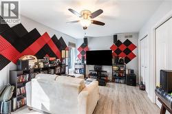 Spacious recreational room featuring wood-finish flooring, built-in shelving and media cabinetry, geometric accent wall with red and black diamond paneling, ceiling fan with integrated lighting, and a - 