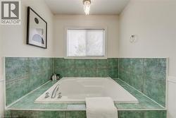 Built-in soaking tub surrounded by green tile, featuring a window with blinds, chrome fixtures, and a crystal chandelier - 