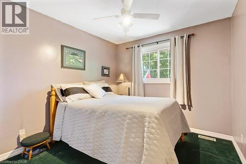 Serene bedroom retreat showcasing soft blush walls, a queen-size bed with quilted bedding, ceiling fan with integrated lighting, large double-hung window with natural light, and dark green-finish floo - 236 Winston Boulevard, Cambridge, ON - Indoor Photo Showing Bedroom