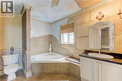 Bathroom featuring a corner soaking tub with tiled surround, a single vanity with a dark countertop and undermount sink, a white toilet, and a window with a woven shade - 74 Forest Edge Trail, Kitchener, ON 