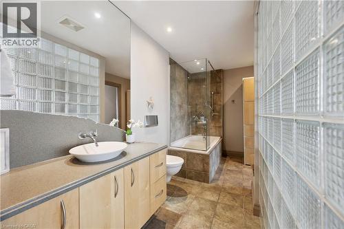 Bathroom featuring a glass block wall, wood-finish cabinetry, a vessel sink, a full-height mirror, and a tiled tub surround - 36 Hayhurst Road Unit# 329, Brantford, ON - Indoor Photo Showing Bathroom