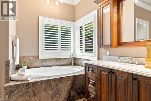 Oval soaking tub, wood-finish vanity with built-in cabinetry, dual-pane windows with white plantation shutters, and a stone-finish tub surround - 253 Rossmore Boulevard, Burlington, ON - Indoor Photo Showing Bathroom
