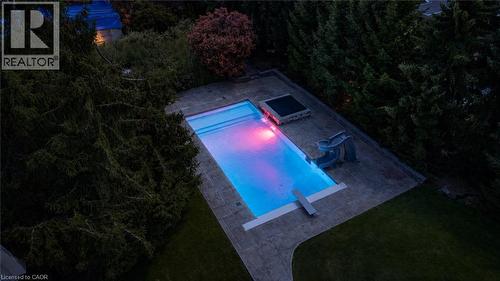 Illuminated resort-style swimming pool with gradient lighting features, complemented by a spacious concrete patio and mature evergreen landscaping - 21 Kerman Avenue, Grimsby, ON 