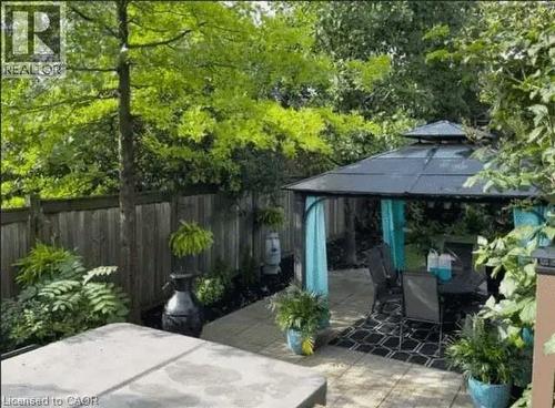 Private patio space featuring a metal-framed gazebo with a dark roof and light blue fabric accents, a paved patio with dark patterned outdoor rug, surrounding lush greenery, and a wooden privacy fence - 4000 Medland Drive, Burlington, ON - Outdoor