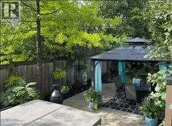Private patio space featuring a metal-framed gazebo with a dark roof and light blue fabric accents, a paved patio with dark patterned outdoor rug, surrounding lush greenery, and a wooden privacy fence - 