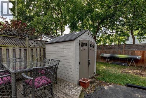 Spacious backyard retreat featuring a gable-roof storage shed with arched doorway and horizontal siding, mature shade trees, wooden deck with integrated seating area, fenced perimeter, and manicured l - 23 Kipling Road, Hamilton, ON - Outdoor With Deck Patio Veranda With Exterior