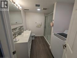 Spacious bathroom featuring a white vanity with integrated sink, glass-enclosed shower, wood-finish flooring, and a modern top-load washer - 