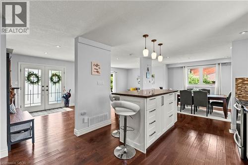 Hardwood-finish flooring, white kitchen island with dark countertop, three pendant lights, dining area with window, and double entry doors with decorative glass inserts - 87 Monte Drive, Hamilton, ON - Indoor