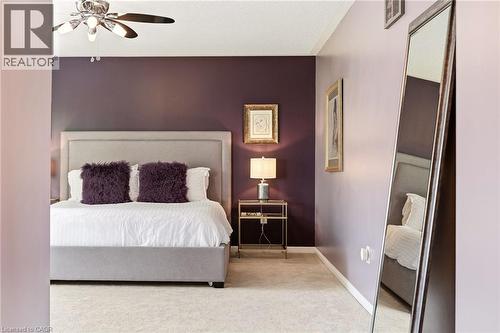 Sophisticated primary bedroom suite featuring a neutral upholstered bed frame, deep plum accent wall, wood-finish flooring, ceiling fan with integrated lighting, and a full-length mirror - 536 Cusick Circle, Milton, ON - Indoor Photo Showing Bedroom