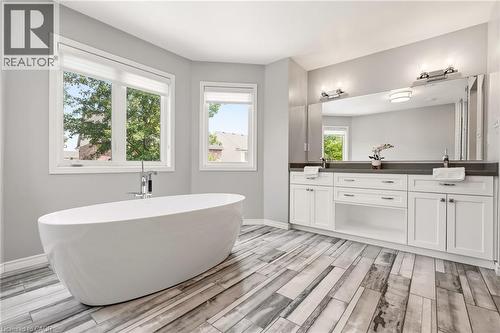 Spa-inspired primary bathroom featuring a freestanding oval soaking tub, dual-sink vanity with white cabinetry and dark countertops, large framed mirror with integrated lighting, wood-finish flooring - 536 Cusick Circle, Milton, ON - Indoor