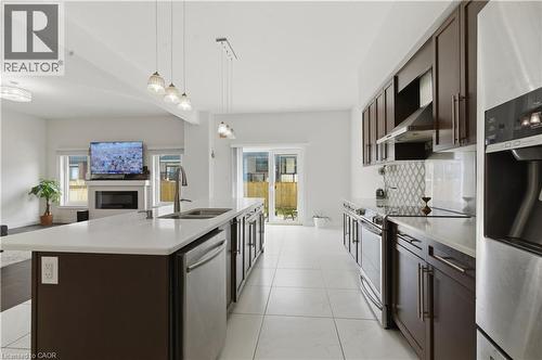 Galley-style kitchen featuring stainless steel appliances, white quartz countertops, dark wood cabinetry with glass-front upper cabinets, pendant lighting over the island, and tile flooring throughout - 2000 Maddex Way, London, ON - Indoor Photo Showing Kitchen With Double Sink With Upgraded Kitchen