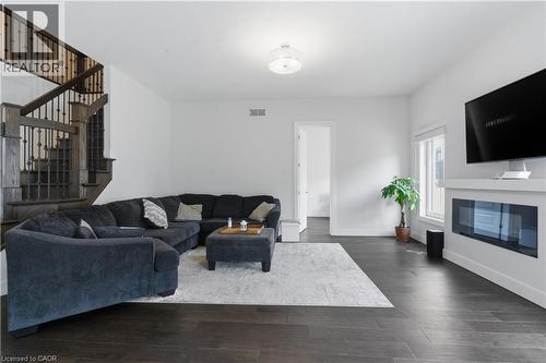 Spacious open-concept living area featuring dark wood-finish flooring, a curved sectional seating arrangement, a linear gas fireplace with white surround, wall-mounted flat-screen television, and abun - 2000 Maddex Way, London, ON - Indoor Photo Showing Living Room With Fireplace