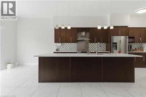 Contemporary kitchen featuring rich espresso cabinetry, stainless steel appliances, a large waterfall-edge island with seating, geometric tile backsplash, pendant lighting, and light wood-finish floor - 2000 Maddex Way, London, ON - Indoor Photo Showing Kitchen With Upgraded Kitchen