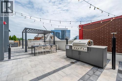 Spacious rooftop patio featuring light-toned paver flooring, a built-in outdoor kitchen with a stainless steel grill, a modern pergola structure, string lighting, and a brick accent wall - 15 Wellington Street S Unit# 903, Kitchener, ON - Outdoor