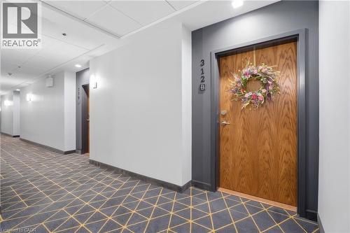 Wood-panel entry door with dark trim, geometric-patterned carpeted hallway, and wall-mounted sconce lighting - 251 Northfield Drive E Unit# 312, Waterloo, ON 